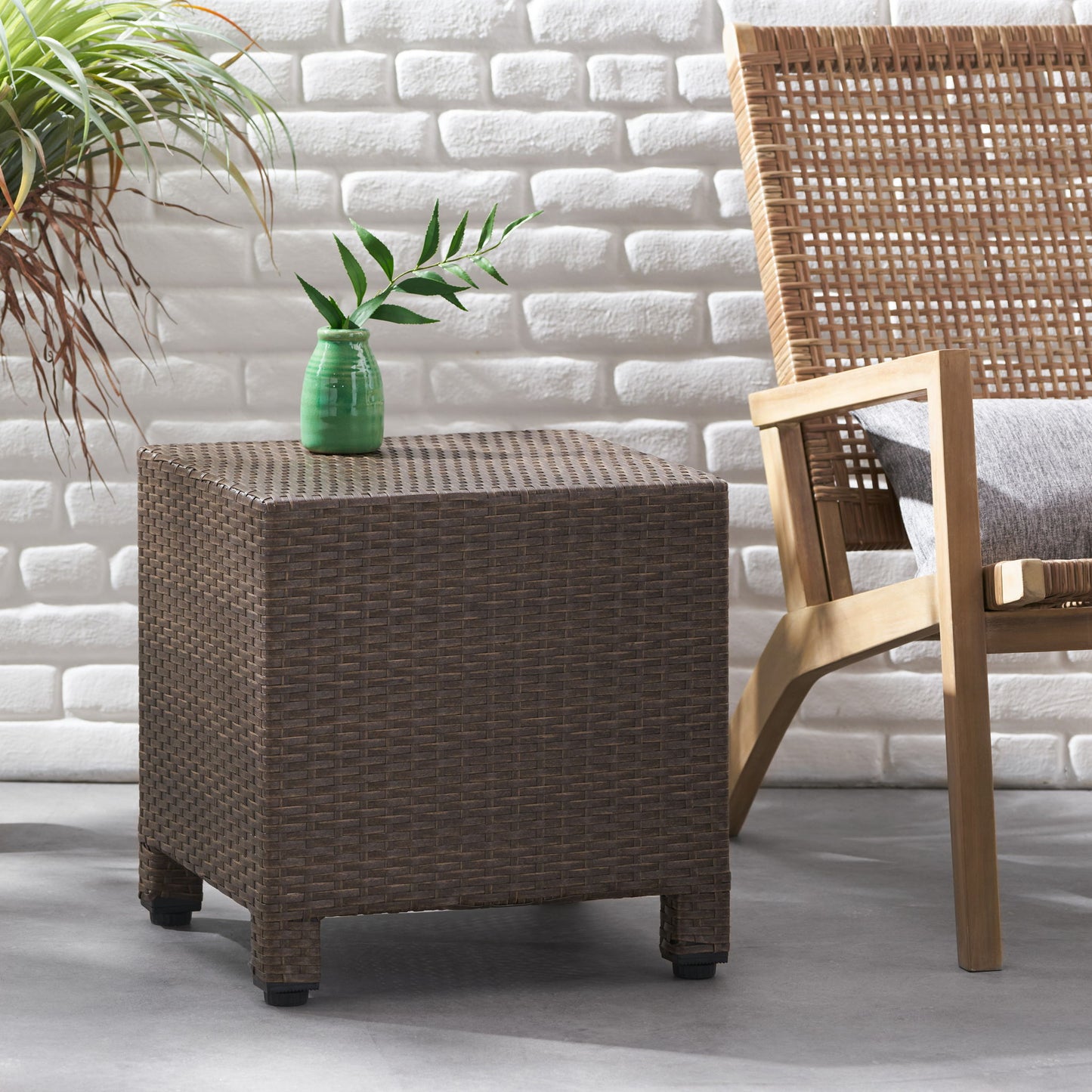 The Puerta - Minimalist Style Accent Table with a small green vase and plant sits beside a wooden chair with a woven back and gray cushion, against a white brick wall and gray floor. A potted plant is on the left.