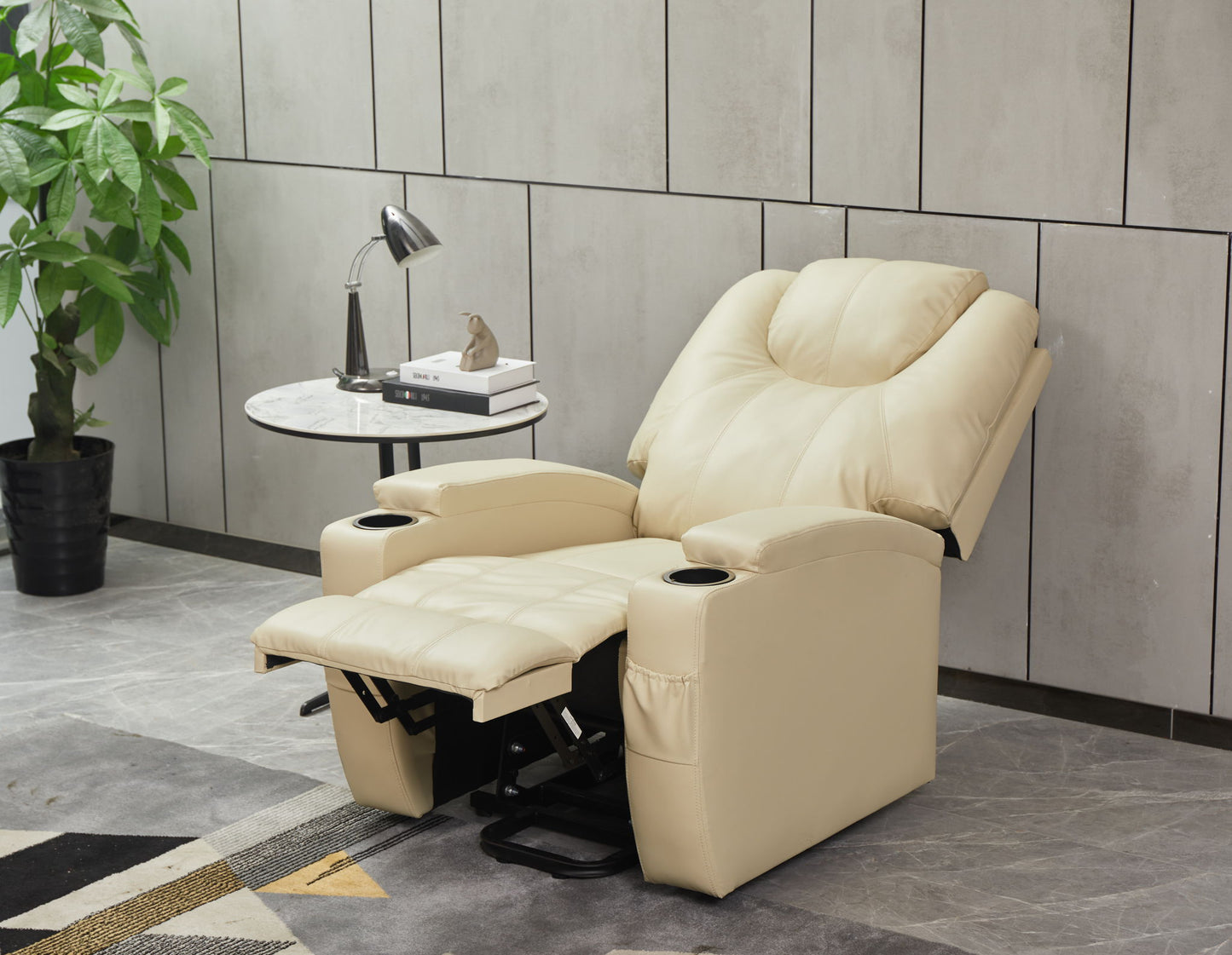 A Modern Recliner Chair With Cup Holders and Remote Control sits by a small round side table featuring a lamp, books, and decor. A potted plant and contemporary tiled wall complete the background.