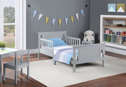 The Connelly Reversible Panel Toddler Bed in modern gray features blue and white bedding. The children's room includes a small gray chair, toy bear on a nightstand, colorful wall bunting, toy shelves, and a scenic window view.