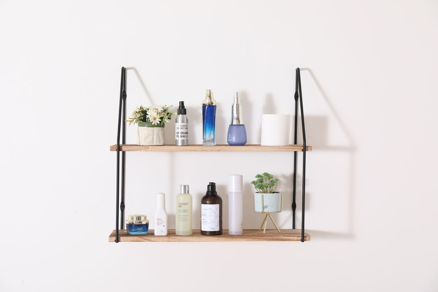 The Gardenia Wall Mounted Wall Storage features black metal supports and two wooden shelves, perfect for displaying skincare bottles, jars, plants, and decorative flowers against a plain white wall.