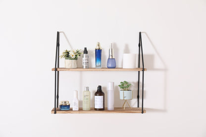 The Gardenia Wall Mounted Wall Storage features black metal supports and two wooden shelves, perfect for displaying skincare bottles, jars, plants, and decorative flowers against a plain white wall.