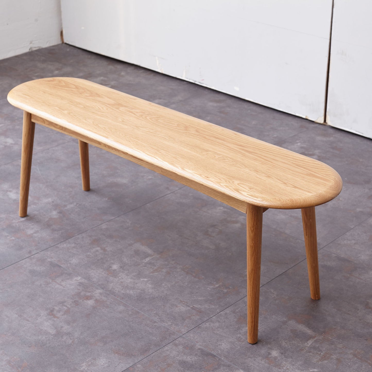 The Dining Bench, Table Bench For Living Room features a simple wooden design with rounded edges and four legs, positioned on a gray tiled floor beside a white wall.