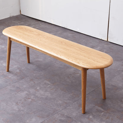 The Dining Bench, Table Bench For Living Room features a simple wooden design with rounded edges and four legs, positioned on a gray tiled floor beside a white wall.