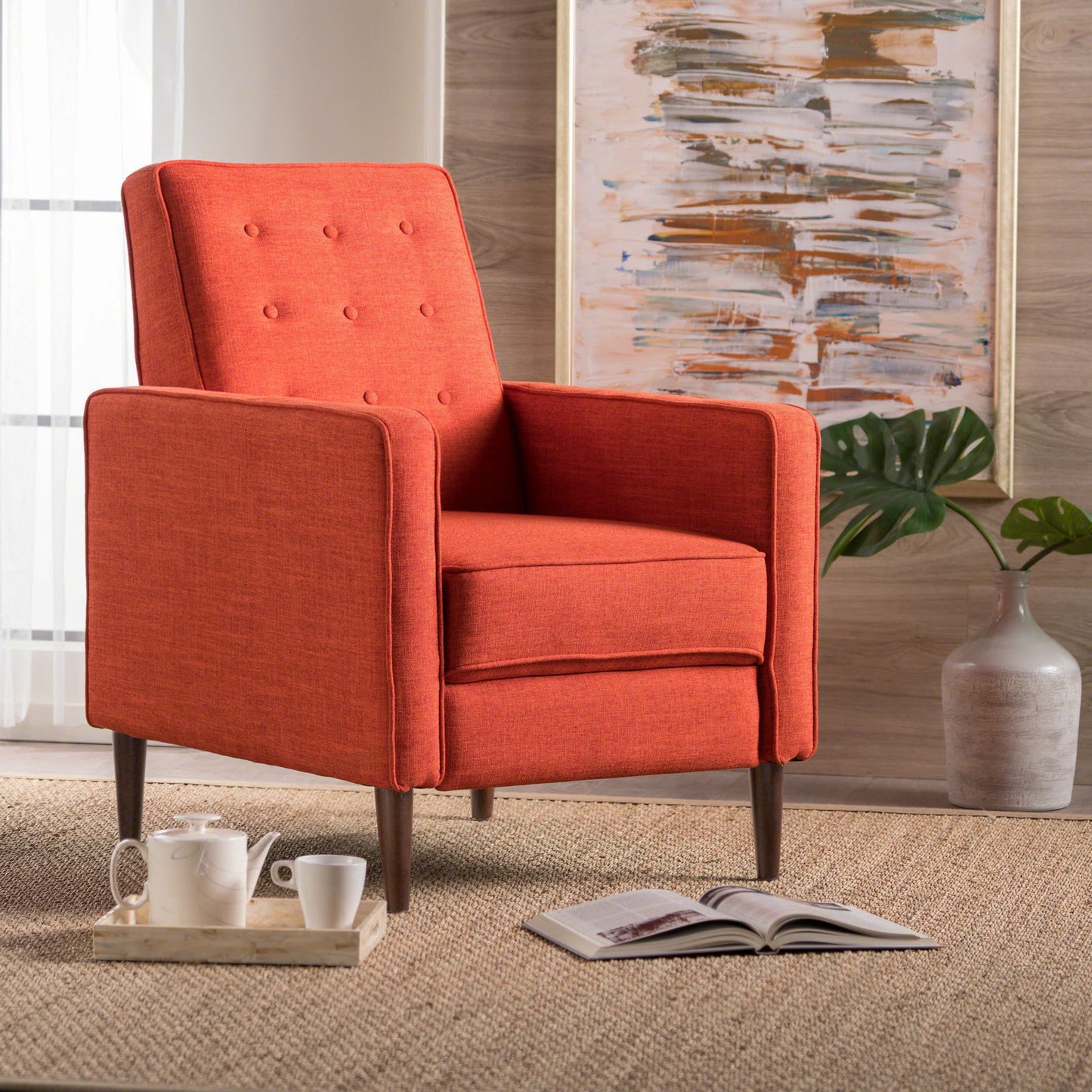 A Mid-Century Modern Recliner With Button Tufted Waffle Stitch in orange sits on a woven rug beside a wooden tray with teacups and an open book, set against a large plant and abstract art painting in the background.