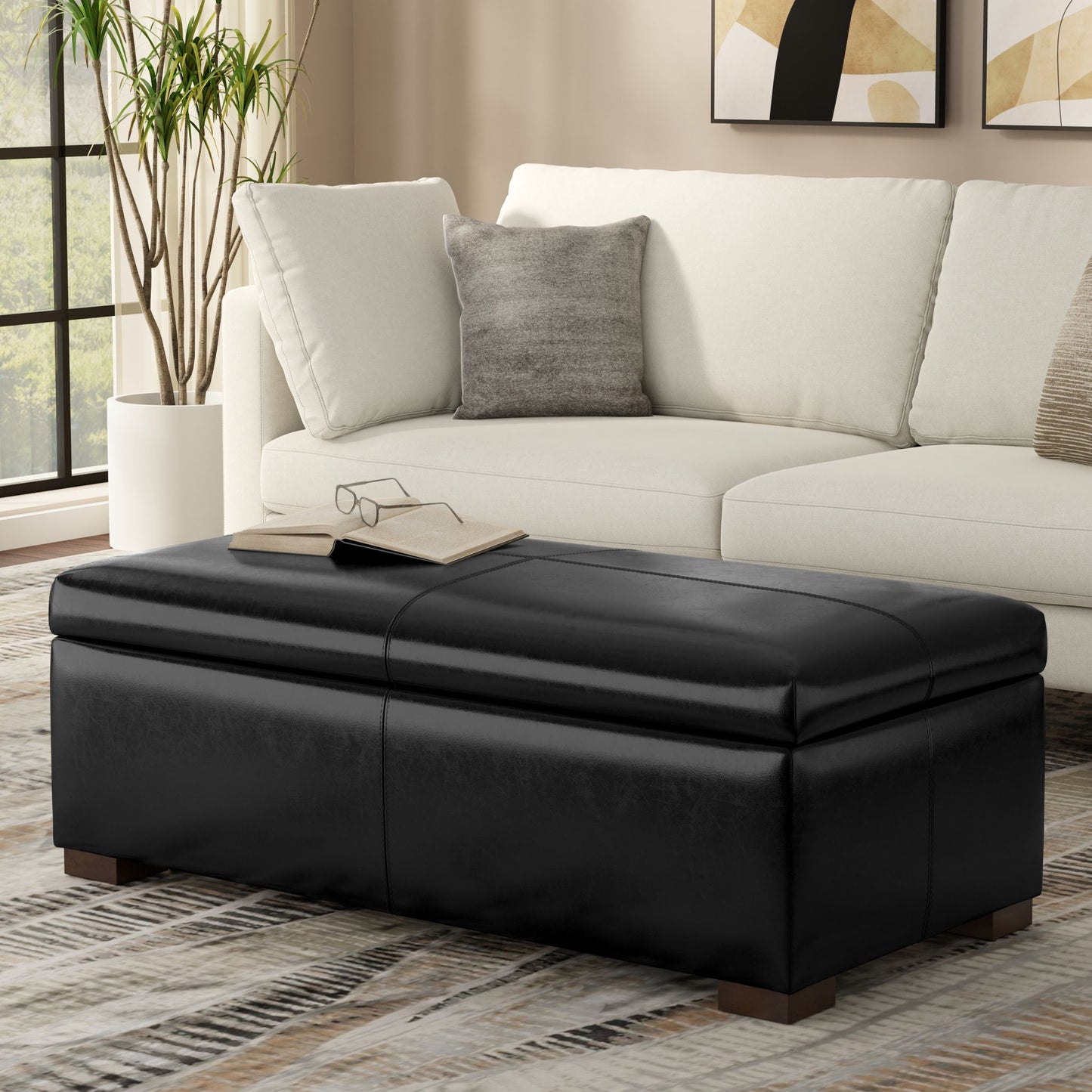 A modern living room with a white sectional sofa, gray pillow, and the Gabbie Coffee Table Upholstered Storage Ottoman topped with a book and glasses. A plant is by the window, and abstract art decorates the wall.