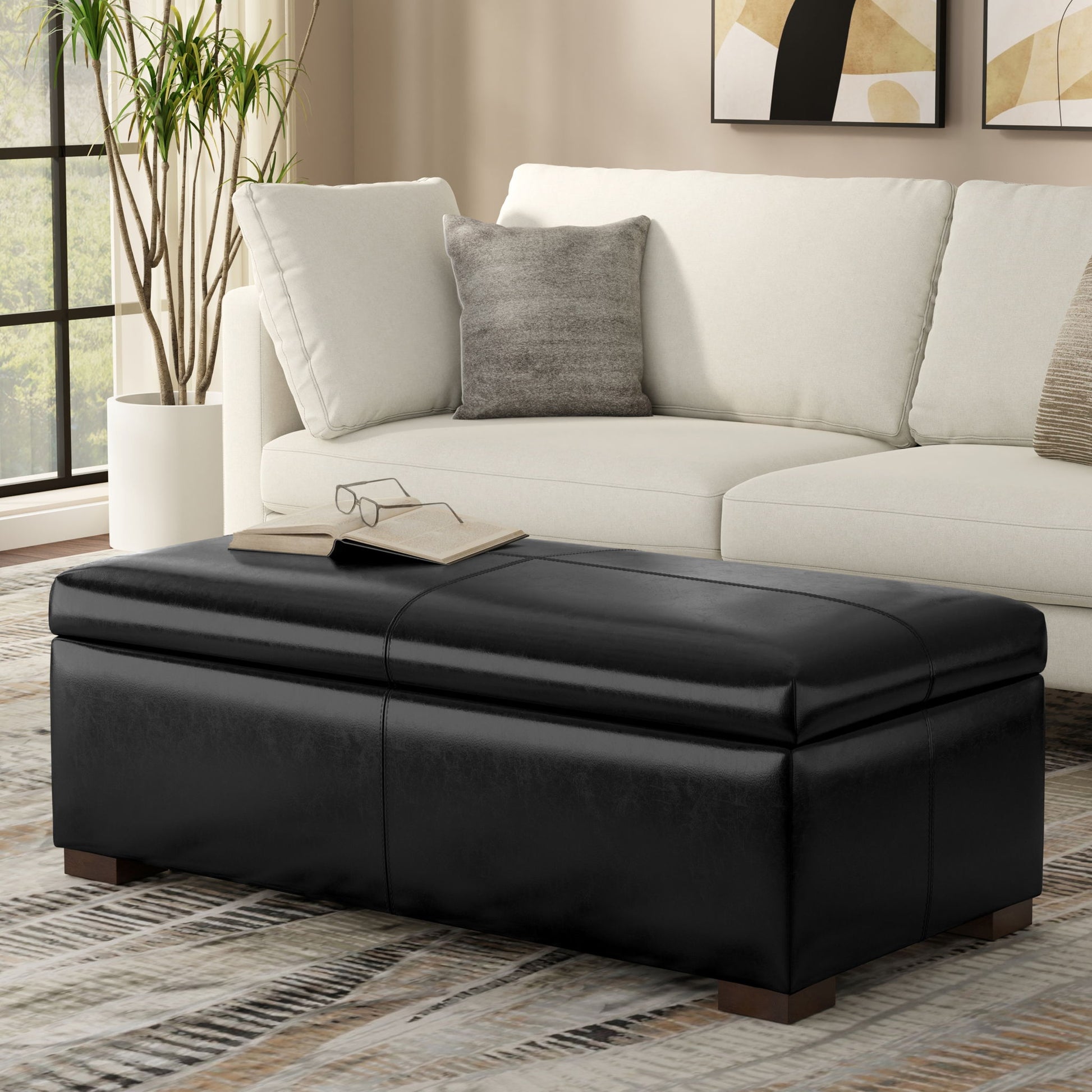A modern living room with a white sectional sofa, gray pillow, and the Gabbie Coffee Table Upholstered Storage Ottoman topped with a book and glasses. A plant is by the window, and abstract art decorates the wall.