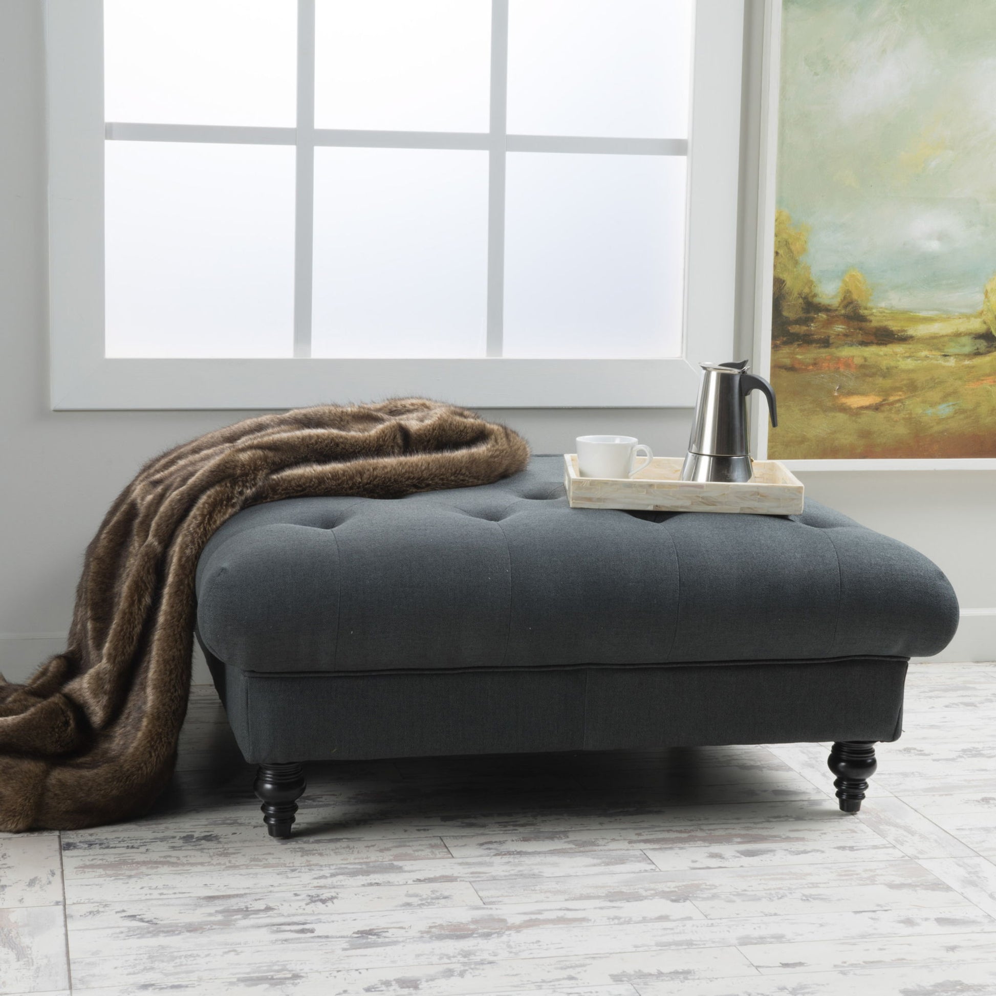The Madrid - Tufted Ottoman Footrest in dark gray rests on a light wood floor by a frosted window, topped with a brown throw blanket and a tray with coffee pot and cup. A landscape painting is displayed on the wall nearby.