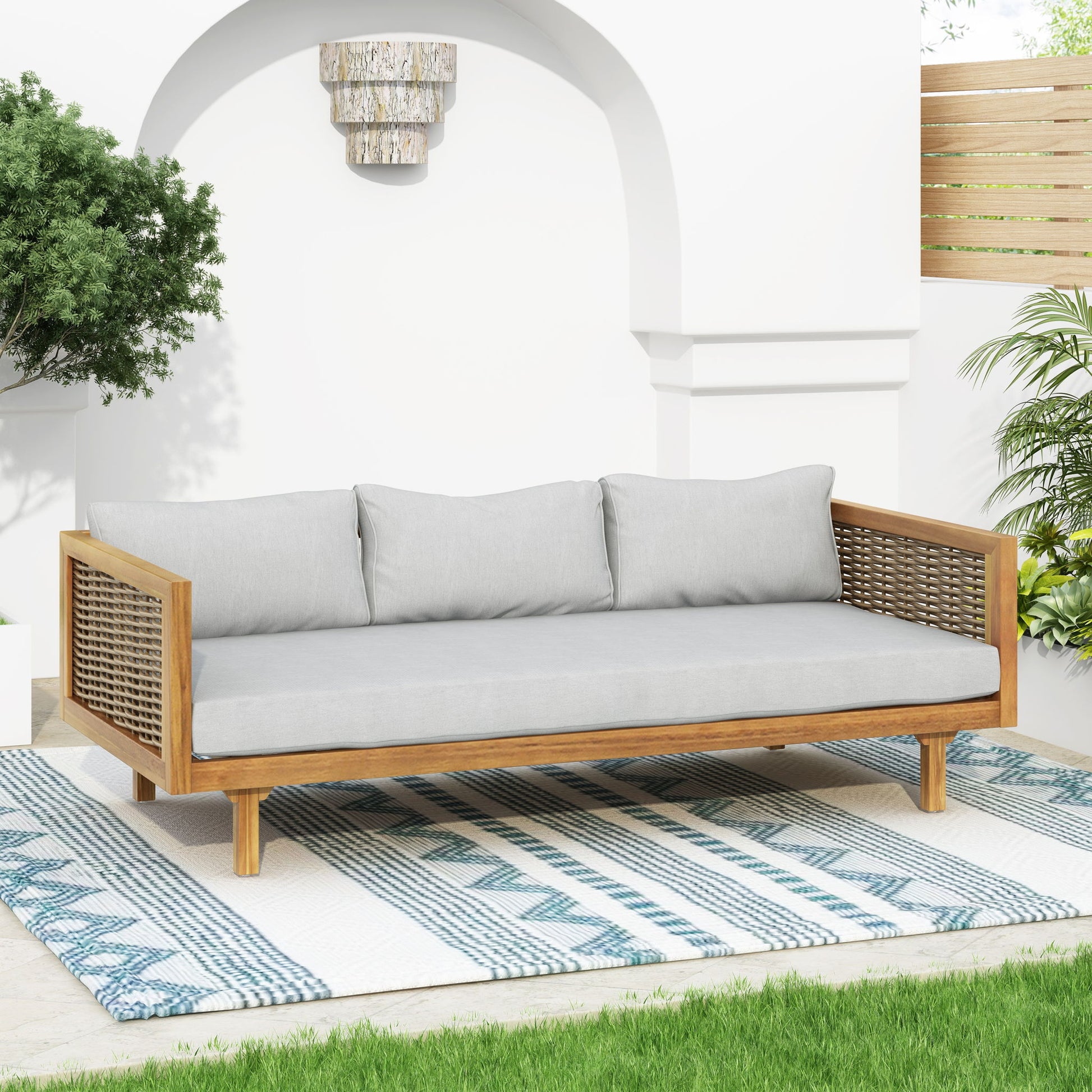 The Claremont 3 Seater Daybed in teak acacia wood with water-resistant light gray cushions is arranged on a blue and white rug, surrounded by green plants, a white wall, and wooden fencing for a modern patio look.