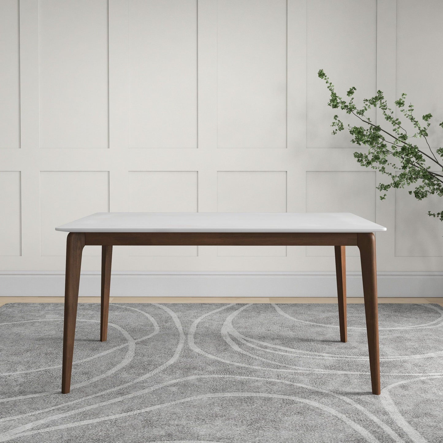The Levi - Modern Style Rectangular Dining Kitchen Table, featuring a white top and dark wooden legs, stands on a gray patterned rug before a white paneled wall, with a vase holding green leaves on the right.