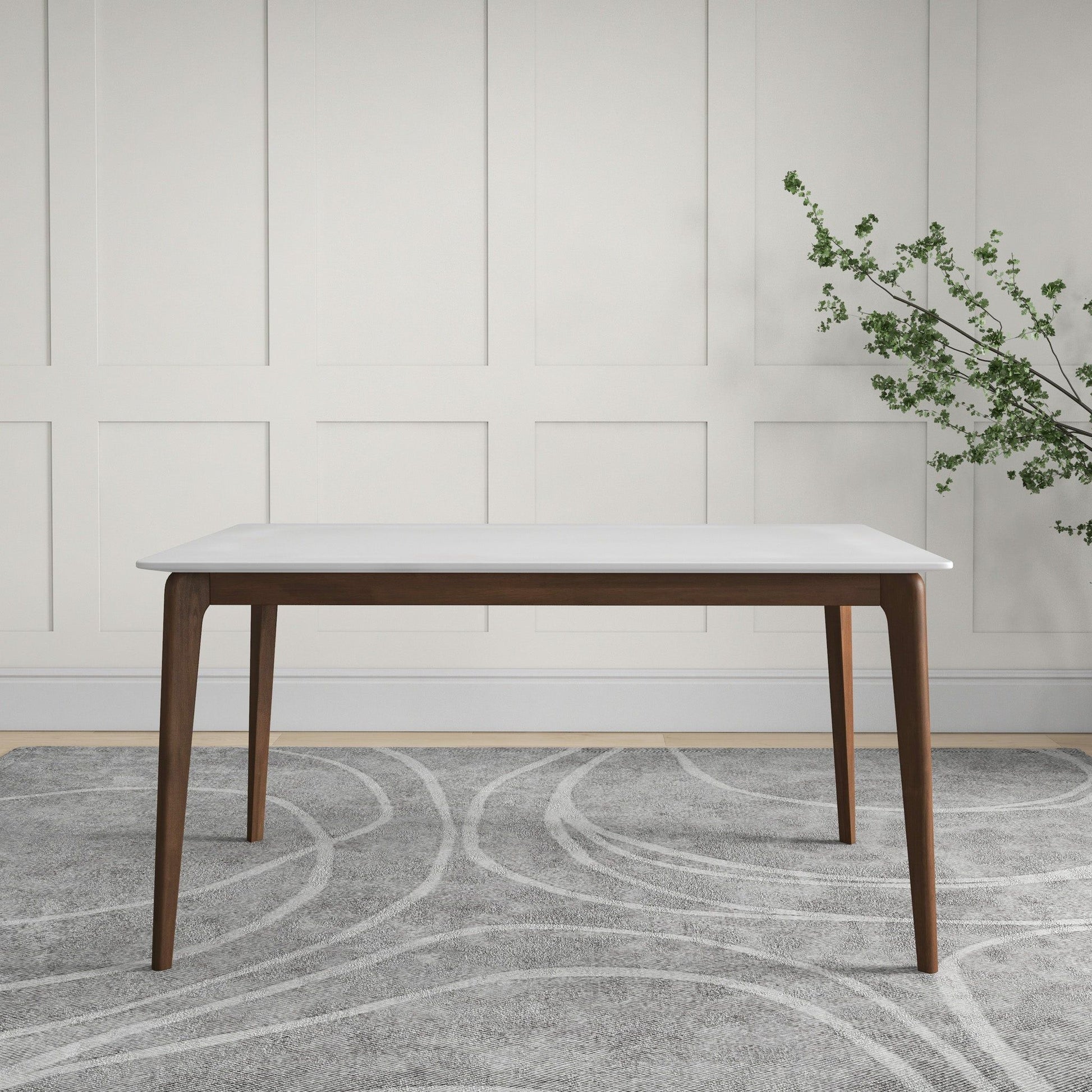 The Levi - Modern Style Rectangular Dining Kitchen Table, featuring a white top and dark wooden legs, stands on a gray patterned rug before a white paneled wall, with a vase holding green leaves on the right.