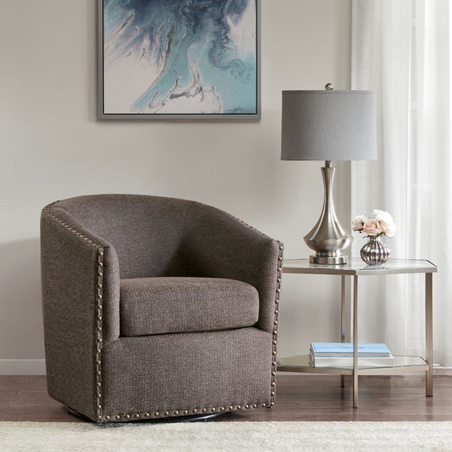 A Luxurious Chair With 360 Degree Rotation in gray upholstery and nailhead trim sits beside a glass-top side table with a silver lamp and flowers. A blue abstract painting hangs above, framed by light curtains.