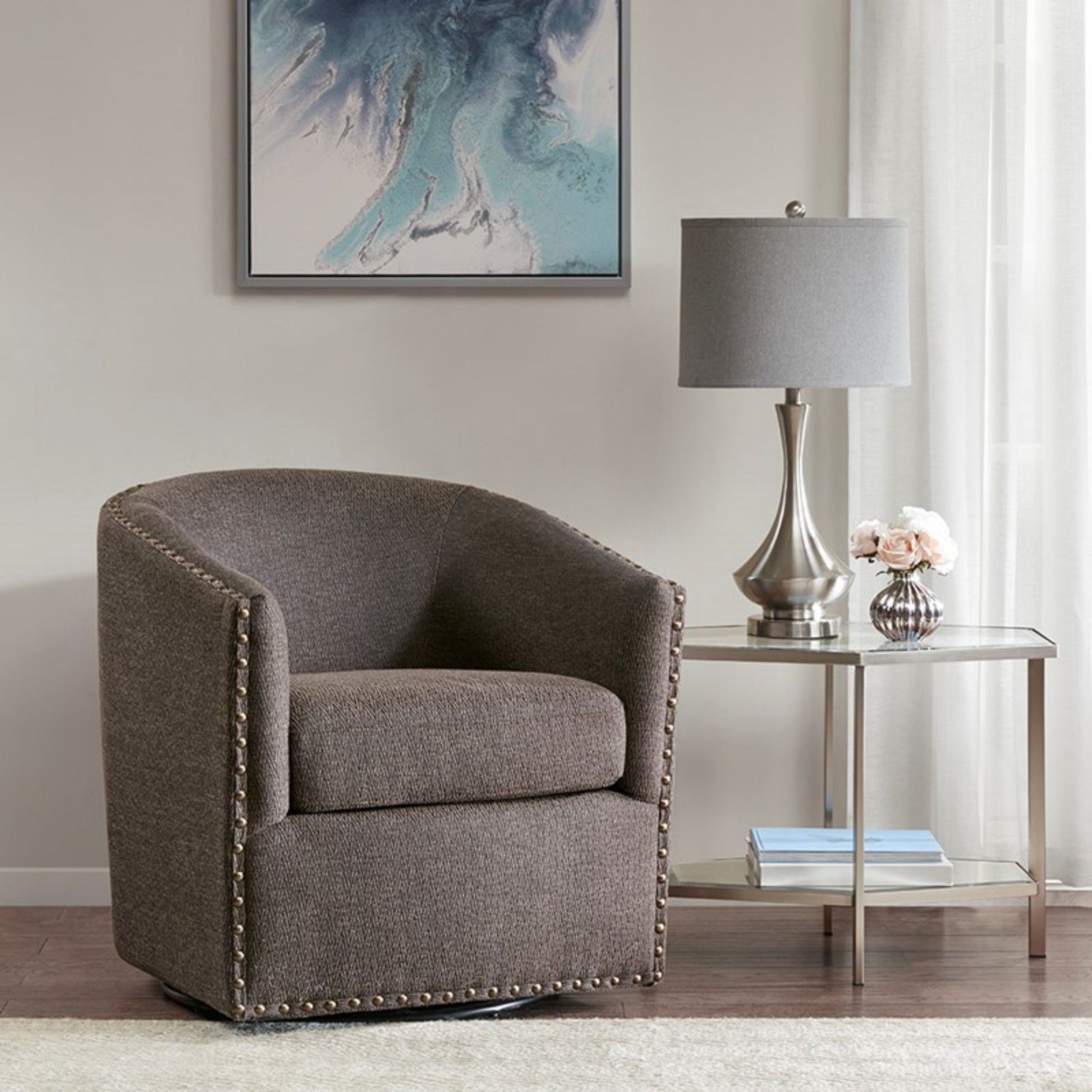 A Luxurious Chair With 360 Degree Rotation in gray upholstery and nailhead trim sits beside a glass-top side table with a silver lamp and flowers. A blue abstract painting hangs above, framed by light curtains.