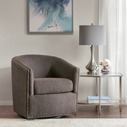 A Luxurious Chair With 360 Degree Rotation in gray upholstery and nailhead trim sits beside a glass-top side table with a silver lamp and flowers. A blue abstract painting hangs above, framed by light curtains.