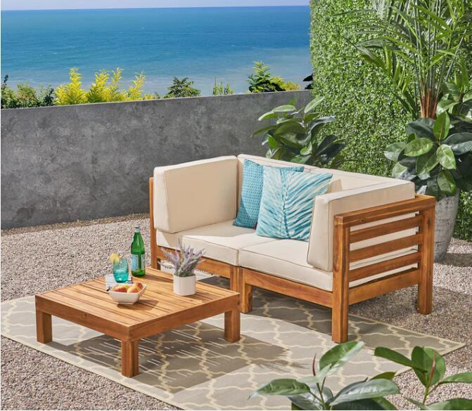 The Oana 3 Piece Love Seat Set with Coffee Table features a wooden outdoor sofa with cream cushions and blue pillows on a patio, alongside a matching coffee table holding drinks and snacks, all framed by ocean views and green plants.