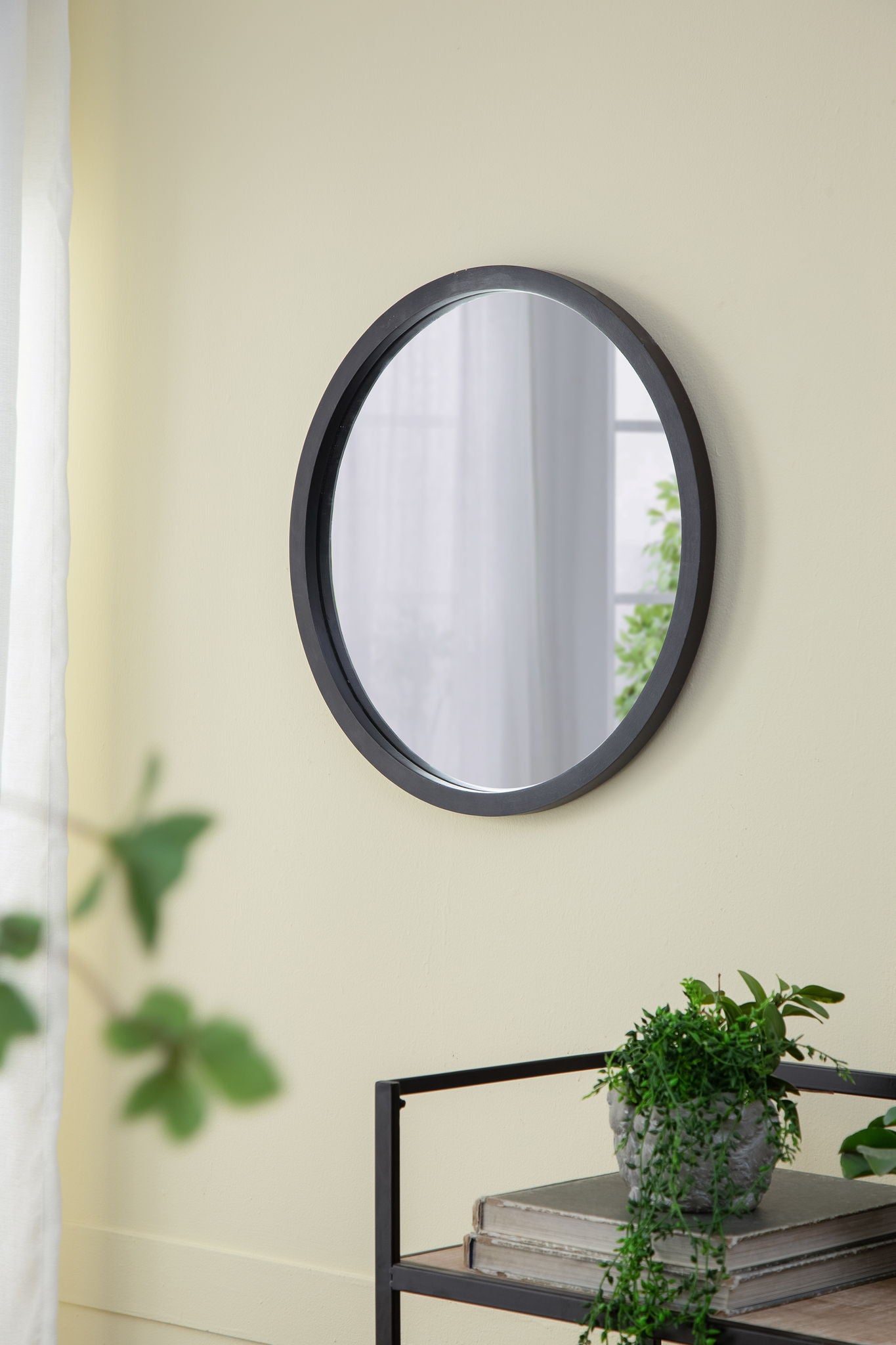 A Circle Wall Mirror With Wooden Frame hangs on a light yellow wall near a window. Beneath it, a black shelf holds potted green plants and stacked books, with leafy plants visible in the foreground.