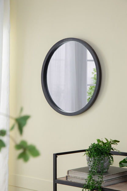 A Circle Wall Mirror With Wooden Frame hangs on a light yellow wall near a window. Beneath it, a black shelf holds potted green plants and stacked books, with leafy plants visible in the foreground.