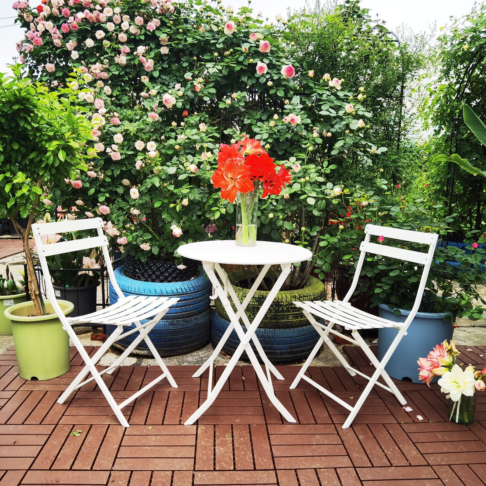 The 3 Piece Patio Bistro Set of Foldable Round Table and Chairs is placed on a wooden patio among green plants and pink roses, with a vase of bright red flowers on the table.