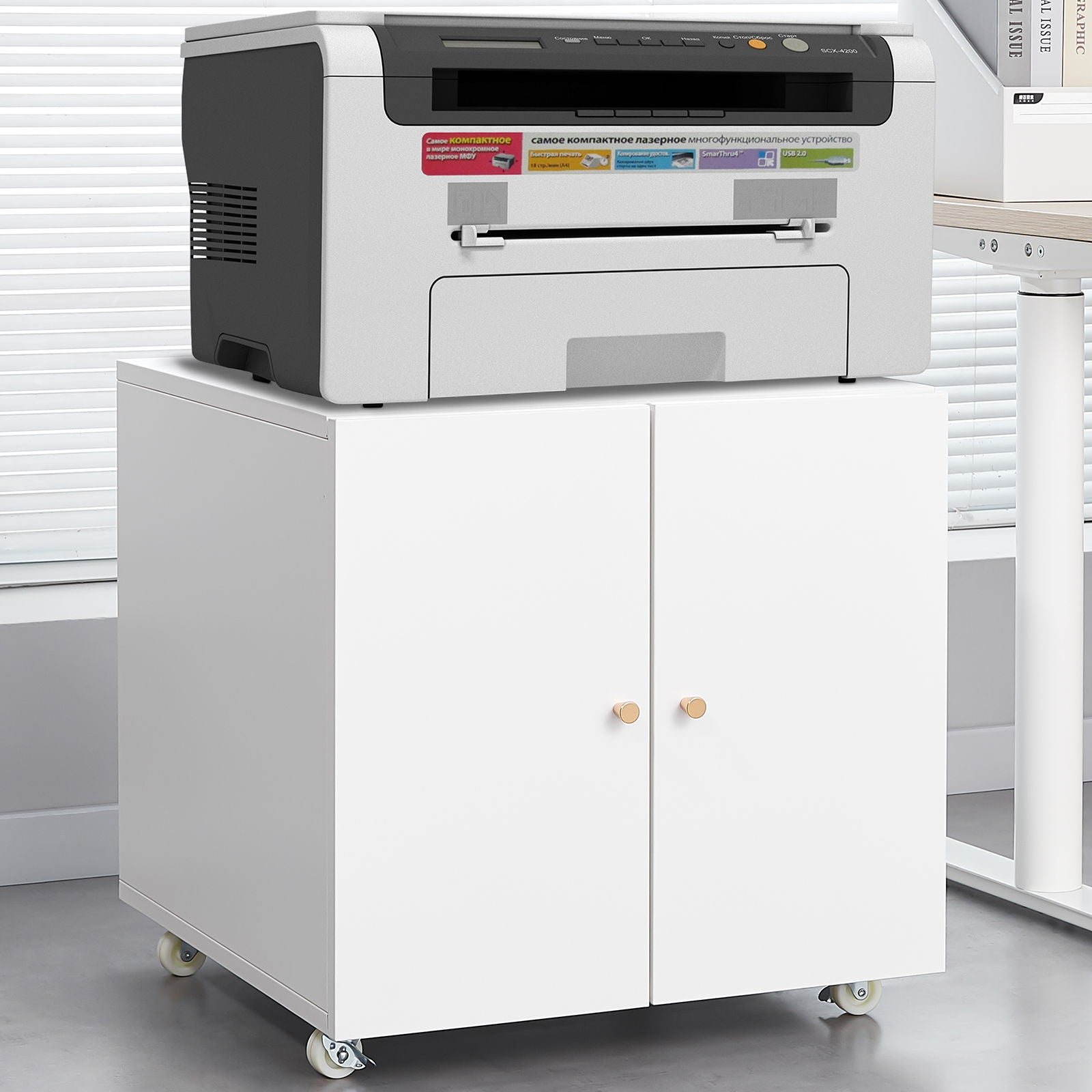 The Office Furniture Copier Cabinet 2 Door Steel Copier Stand Mobile Pedestal File Printer Stand holds a gray printer and features two doors with wooden knobs, wheels for mobility, and fits well in an office with blinds and a gray floor.