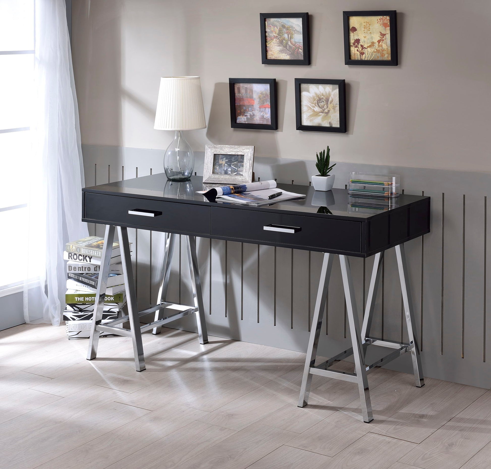 The Coleen High Gloss Writing Desk With USB in black with chrome legs sits by a light gray wall, adorned with framed art, a white lamp, books, photo frame, and small potted plant on a light wood floor near a window with sheer curtains.