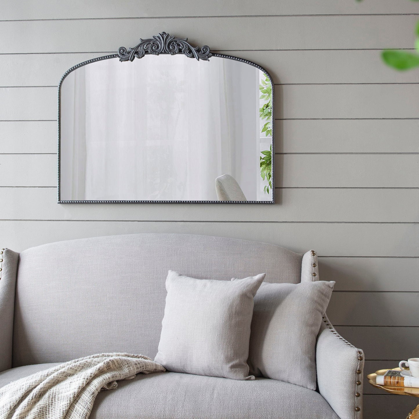 The Arch Mirror, Baroque Inspired Wall Decor, hangs on a light gray shiplap wall above a gray upholstered sofa with matching pillows and a throw blanket. A touch of greenery is reflected in the mirror.