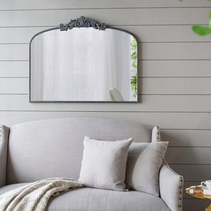 The Arch Mirror, Baroque Inspired Wall Decor, hangs on a light gray shiplap wall above a gray upholstered sofa with matching pillows and a throw blanket. A touch of greenery is reflected in the mirror.