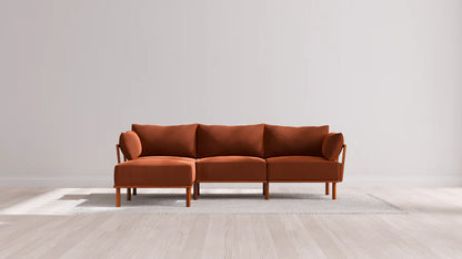 A modern brown sectional sofa with wooden legs is placed on a light gray rug on a wooden floor. The background features a plain white wall, emphasizing the minimalist design of the room.