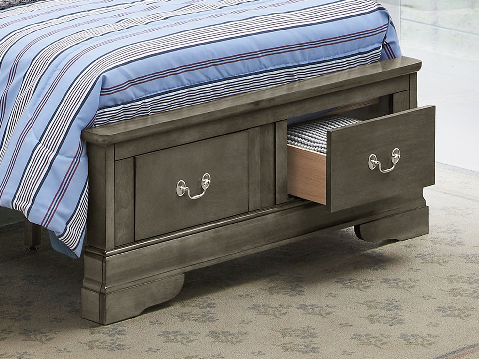 The Louis Phillipe - Storage Bed features two built-in storage drawers at the foot, with one partially open to reveal folded clothes. The bed is topped with a blue striped comforter.