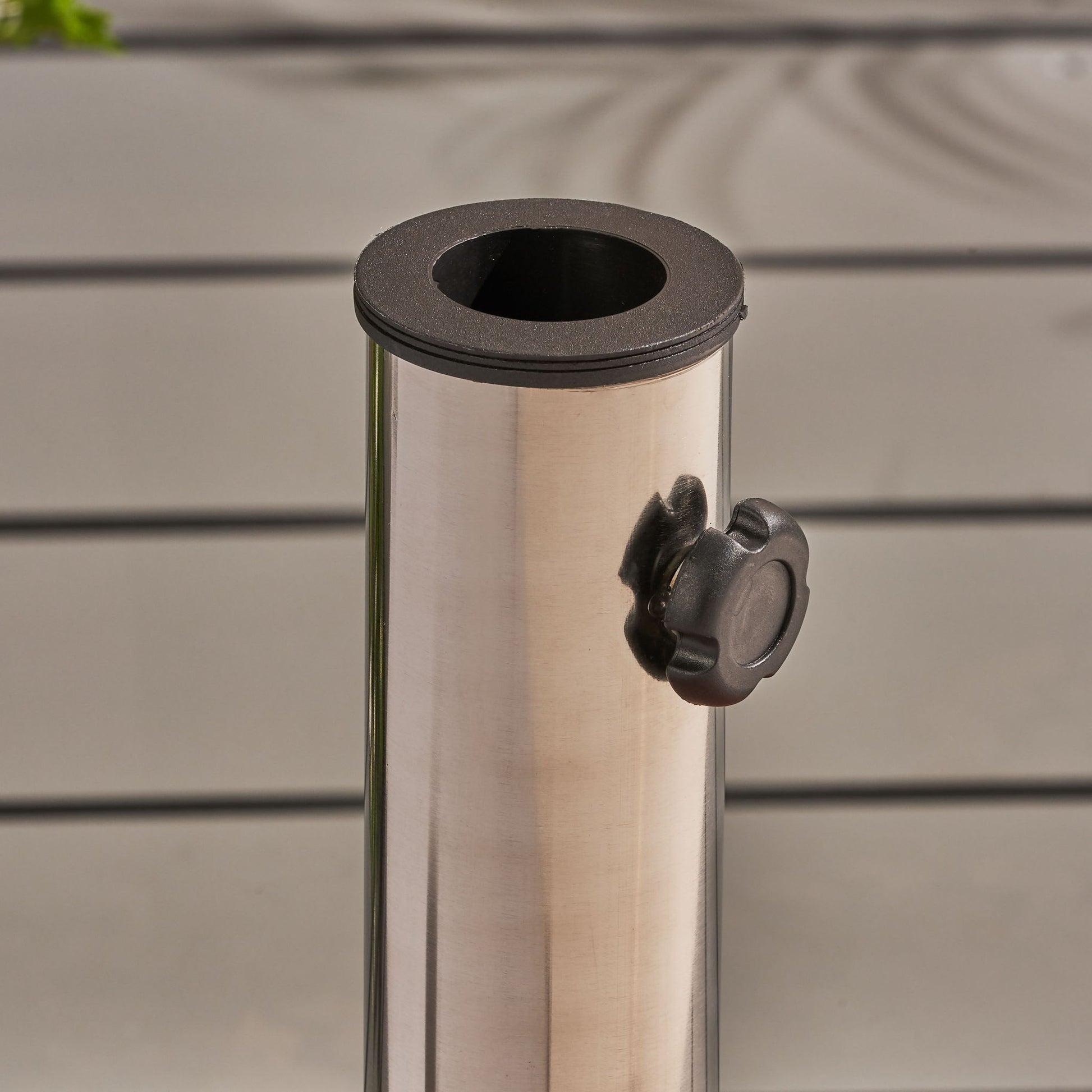 A close-up of the Wheelie Round Umbrella Base with a stainless steel holder and black tightening knob, set against light gray slatted panels.