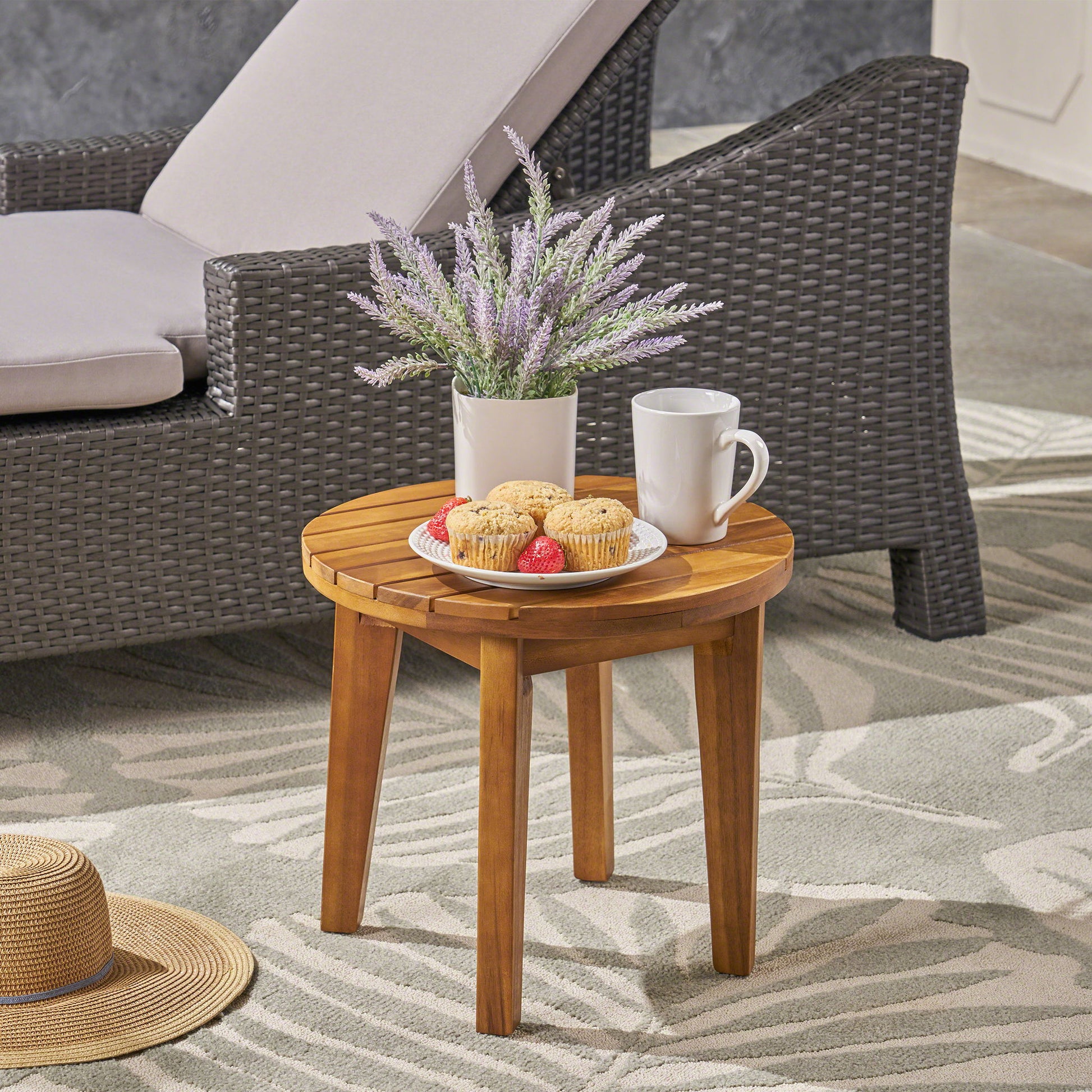 An Outdoor Acacia Wood Side Table With Weather Resistance holds cookies with strawberries, a white mug, and a potted lavender plant. A wicker chair and sun hat rest nearby on a patterned rug.