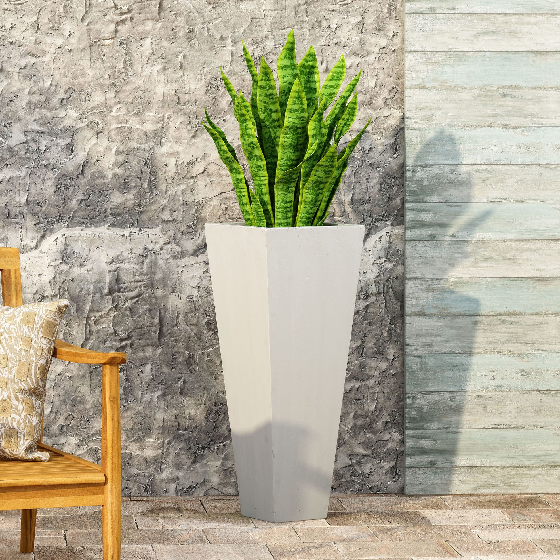 An Outdoor Modern Cast Stone Planter with a snake plant sits on a stone patio next to a textured gray wall and a wooden chair with a patterned cushion.