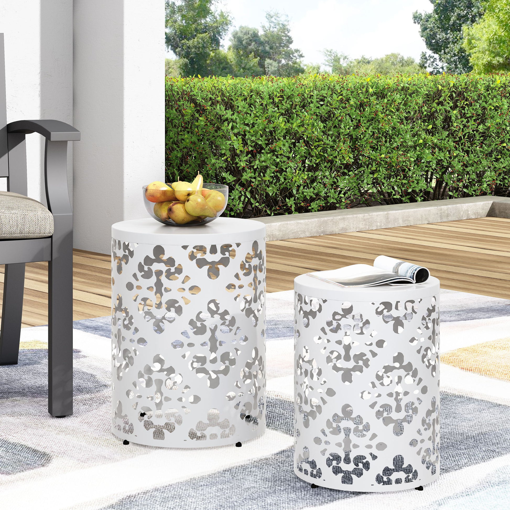 Two white Outdoor Metal Modern Design End Tables with cut-out patterns sit on a patio rug; one holds a bowl of fruit, the other a rolled magazine. Green bushes and a wooden deck are visible in the background.