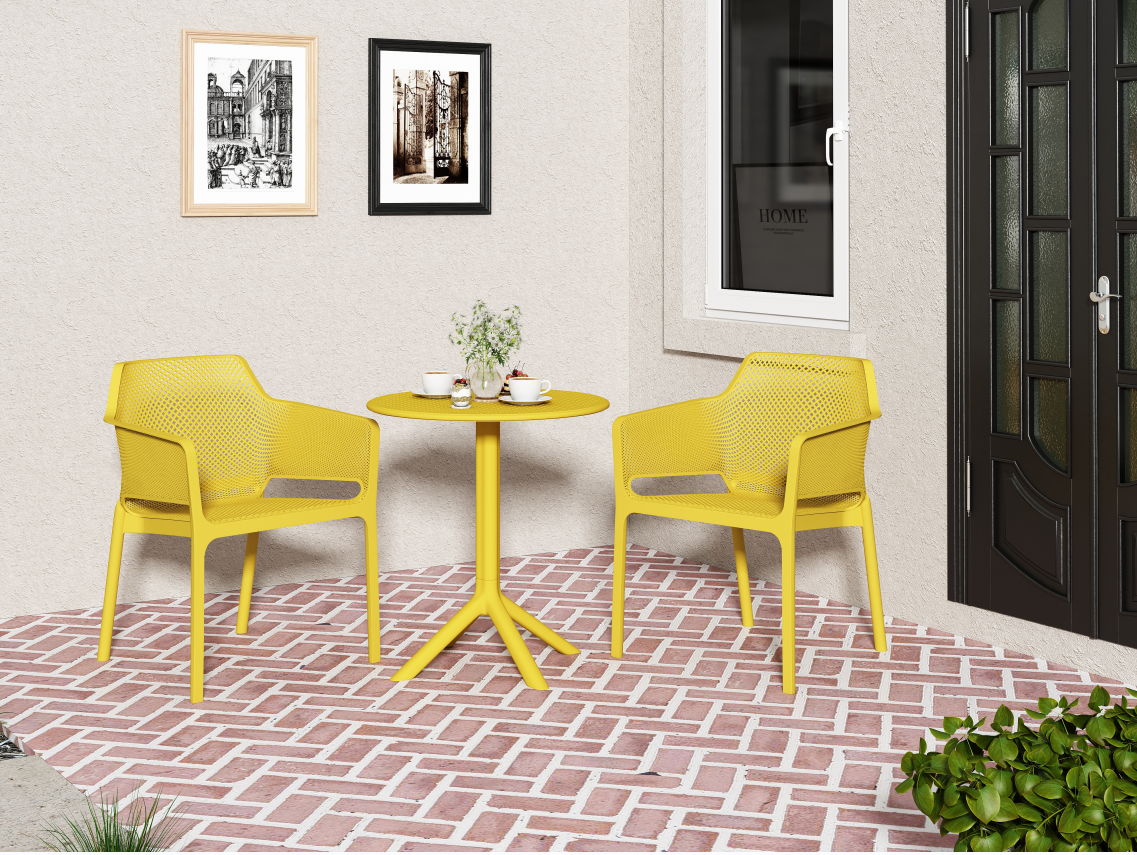 A small patio features two yellow 3 Piece Plastic Arm Chair Bistro Grs Premium Ocean Plastic chairs and a matching round table on red herringbone brick flooring, with cups and a vase on the table, framed pictures on the wall, plus a window and door nearby.
