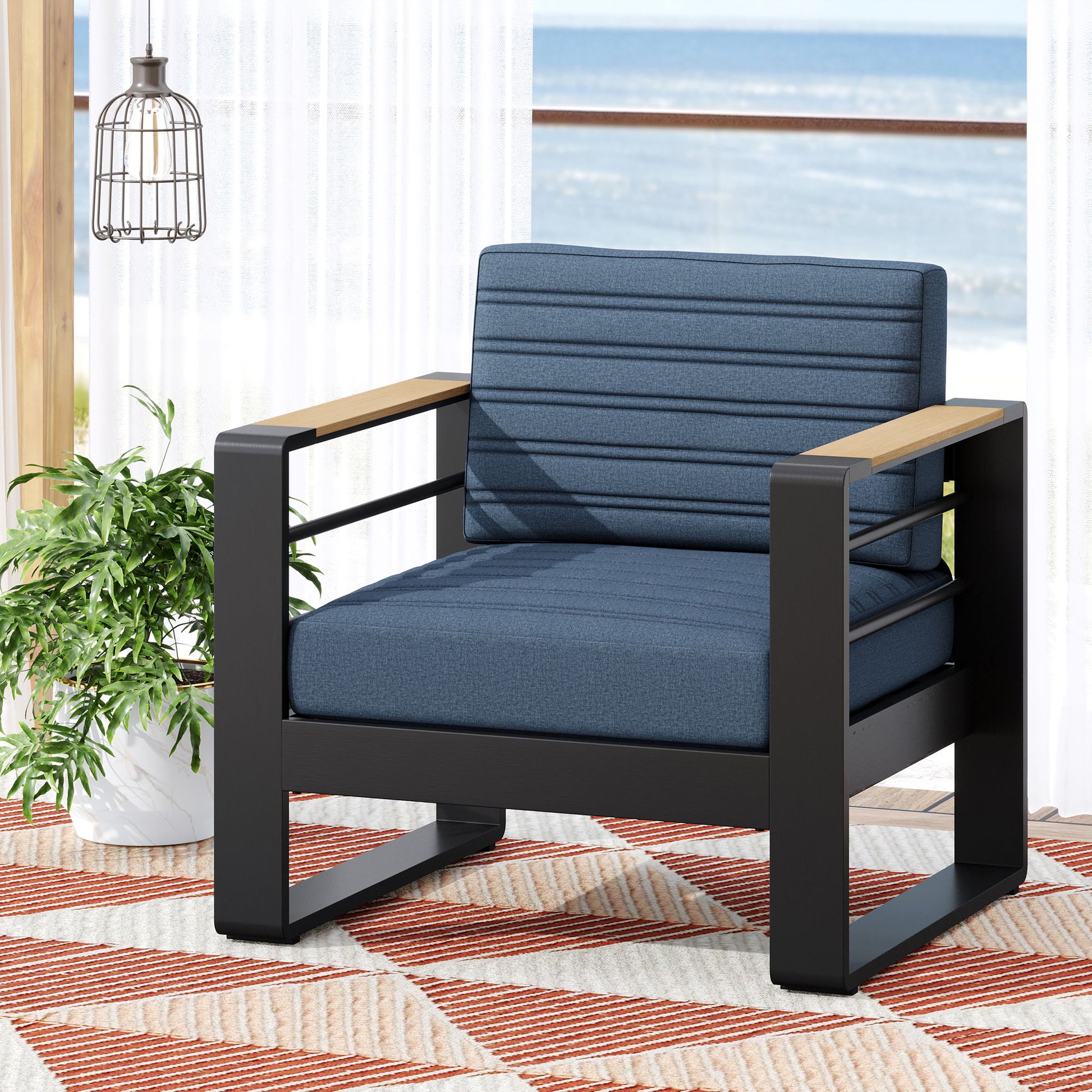 The Giovanna Club Chair Set features a black aluminum frame, blue cushions, and wooden armrests. It sits on a patterned rug beside a green plant, with sheer curtains and an ocean view in the background.