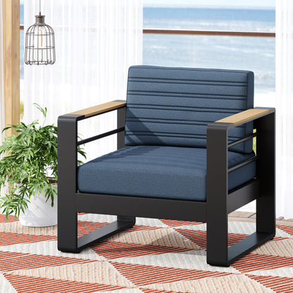 The Giovanna Club Chair Set features a black aluminum frame, blue cushions, and wooden armrests. It sits on a patterned rug beside a green plant, with sheer curtains and an ocean view in the background.