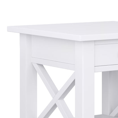 Close-up of the Kitchener - Handcrafted Table, a modern white wooden side table featuring a drawer and X-shaped cross-brace sides, set against a plain white background.