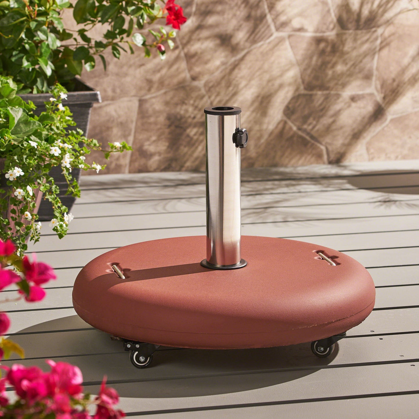 The Wheelie - Round Umbrella Base With Stainless Steel Holder stands on a light gray deck, its reddish-brown form and wheels surrounded by potted green plants and vibrant flowers.