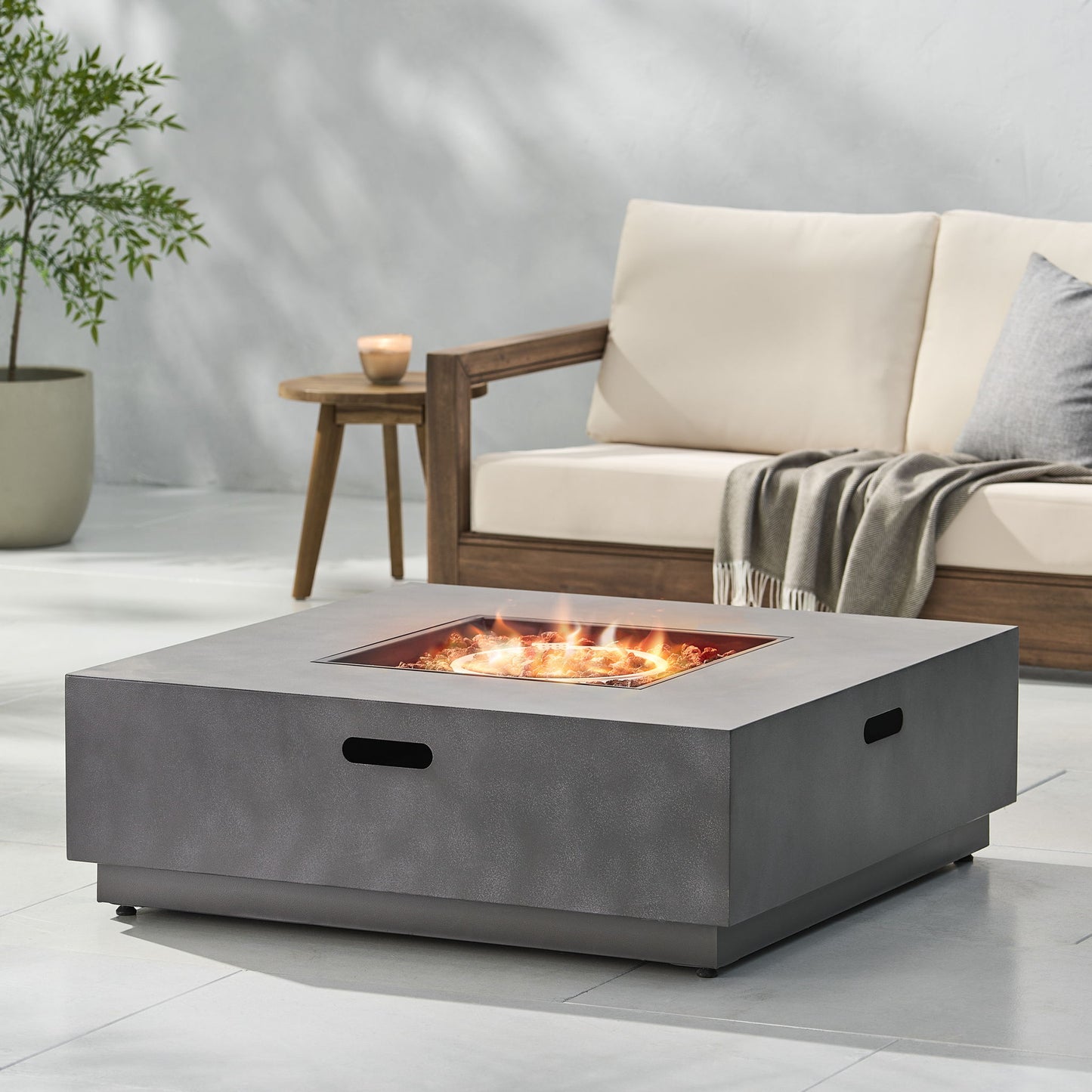 A modern patio features the Modern Square Fire Pit With 50,000 Btu burning brightly, a cushioned wooden sofa with a gray throw, a small wooden side table with a candle, and a potted plant in the background.