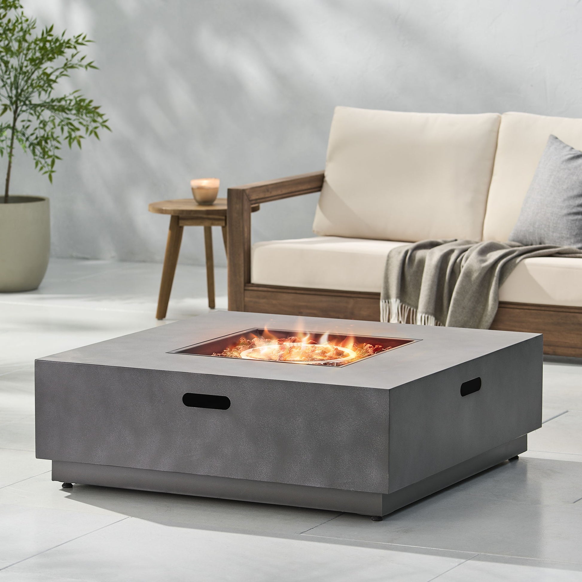 A modern patio features the Modern Square Fire Pit With 50,000 Btu burning brightly, a cushioned wooden sofa with a gray throw, a small wooden side table with a candle, and a potted plant in the background.