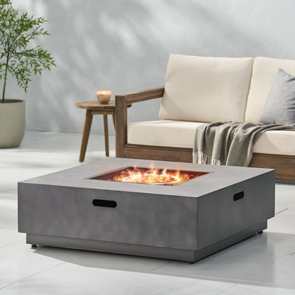 A modern patio features the Modern Square Fire Pit With 50,000 Btu burning brightly, a cushioned wooden sofa with a gray throw, a small wooden side table with a candle, and a potted plant in the background.