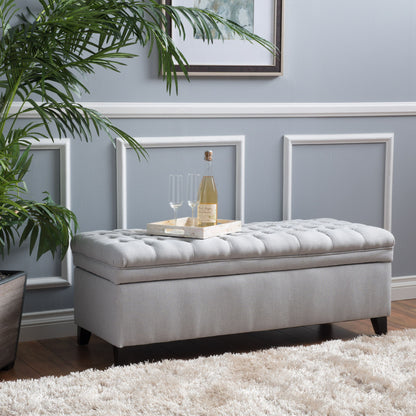 The Juliana Storage Ottoman with Birch Legs in light gray sits on a white rug by a gray wall with white trim. A tray with wine and glasses rests on top, next to a potted plant and framed art.