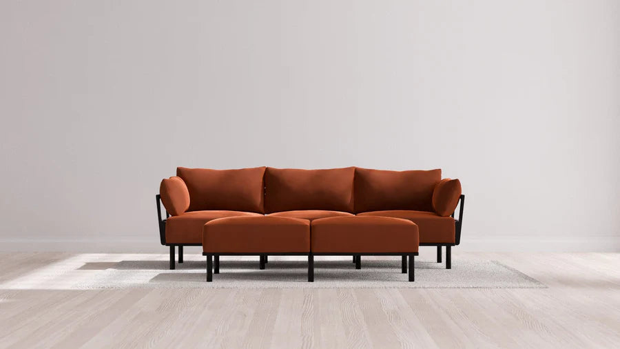 A modern living room features the Indoor Jack 3-Seater Sofa and Bench Ottoman in Balsa, showcasing a vibrant orange upholstery with black legs and armrests. The stylish set is elegantly placed on a light gray rug, framed by a plain white wall and wooden flooring.