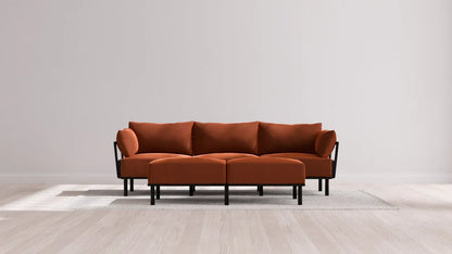 A modern living room features the Indoor Jack 3-Seater Sofa and Bench Ottoman in Balsa, showcasing a vibrant orange upholstery with black legs and armrests. The stylish set is elegantly placed on a light gray rug, framed by a plain white wall and wooden flooring.