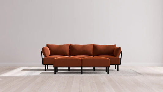 A modern living room features the Indoor Jack 3-Seater Sofa and Bench Ottoman in Balsa, showcasing a vibrant orange upholstery with black legs and armrests. The stylish set is elegantly placed on a light gray rug, framed by a plain white wall and wooden flooring.