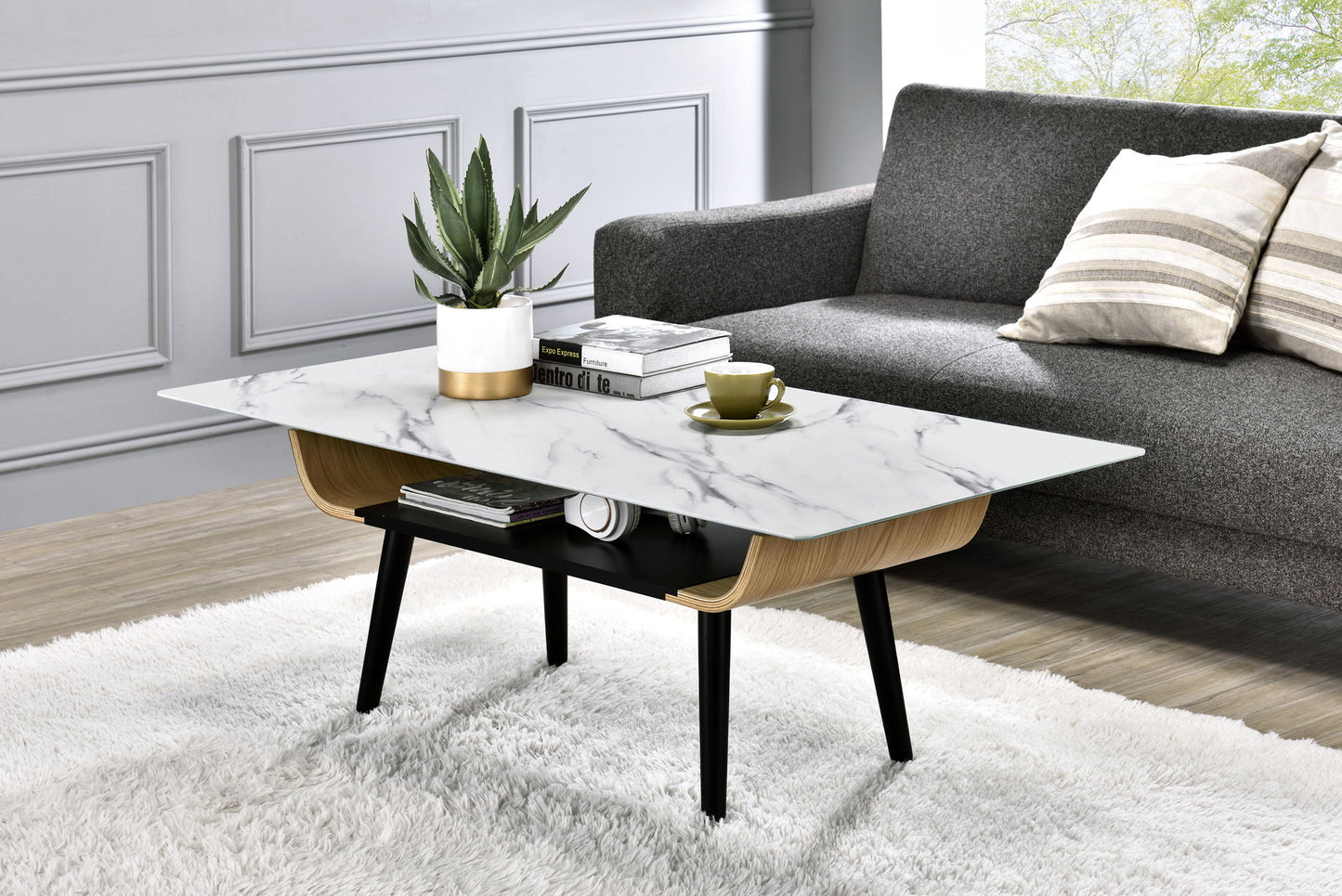 A modern living room features the Landon Coffee Table with a glass marble texture top and bent wood design, displaying a potted plant, books, and a green teacup. The table rests on a white rug before a gray sofa with striped pillows and sunlight streaming in.