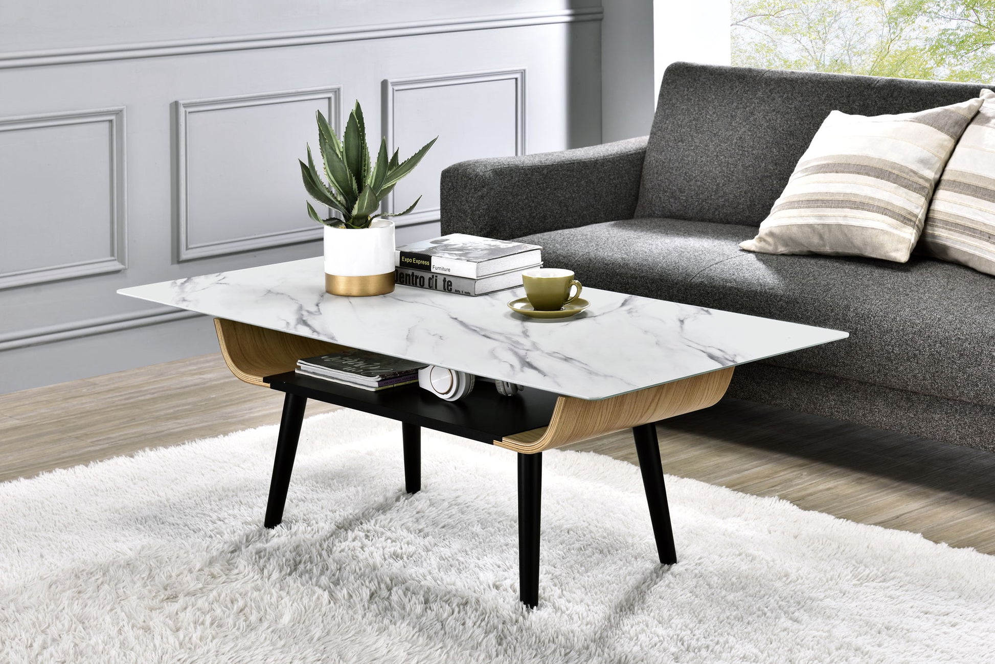 A modern living room features the Landon Coffee Table with a glass marble texture top and bent wood design, displaying a potted plant, books, and a green teacup. The table rests on a white rug before a gray sofa with striped pillows and sunlight streaming in.