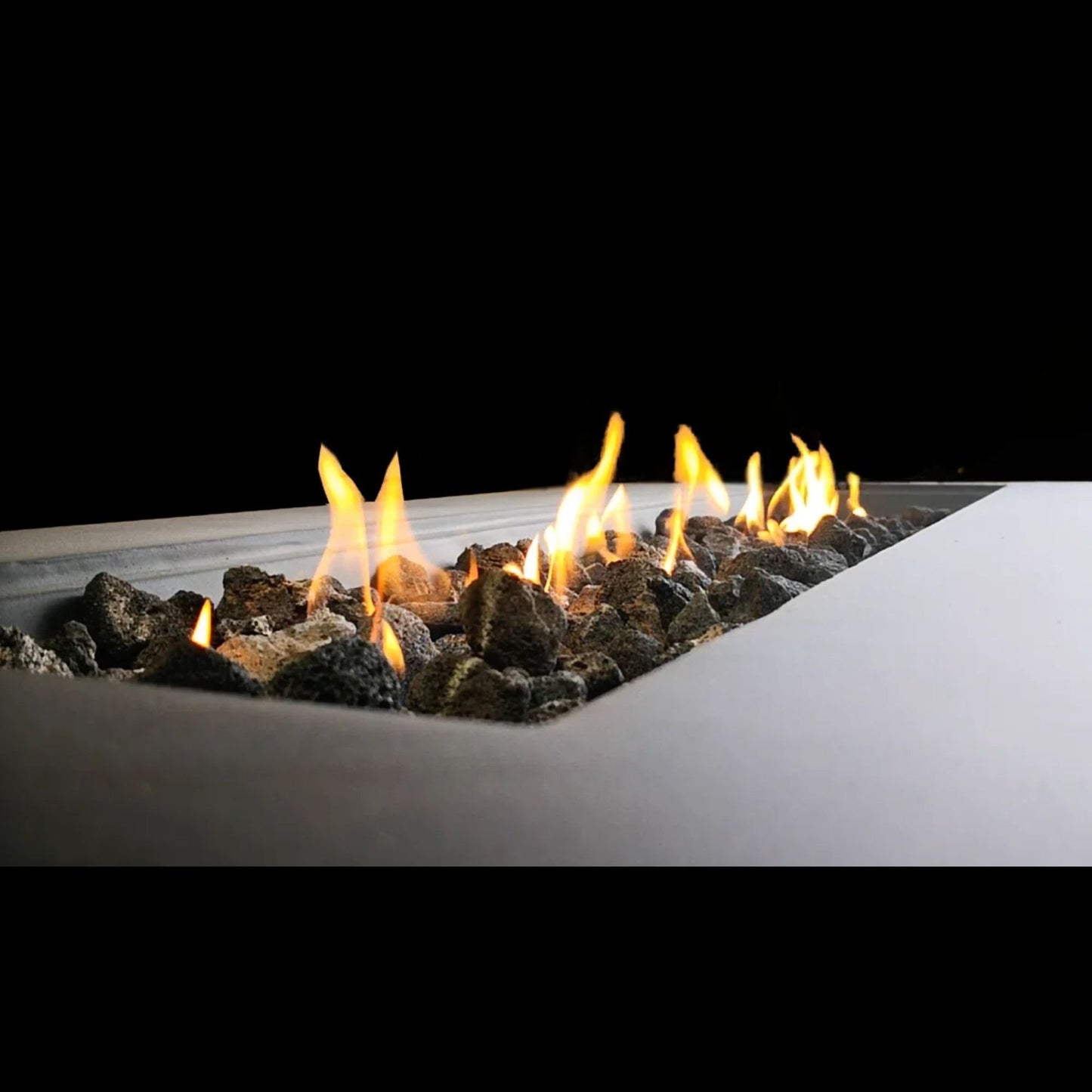 Modern Concrete Design Propane Outdoor Fire Pit Table features bright yellow flames rising from the center of dark stones in a rectangular pit, set against a black background.
