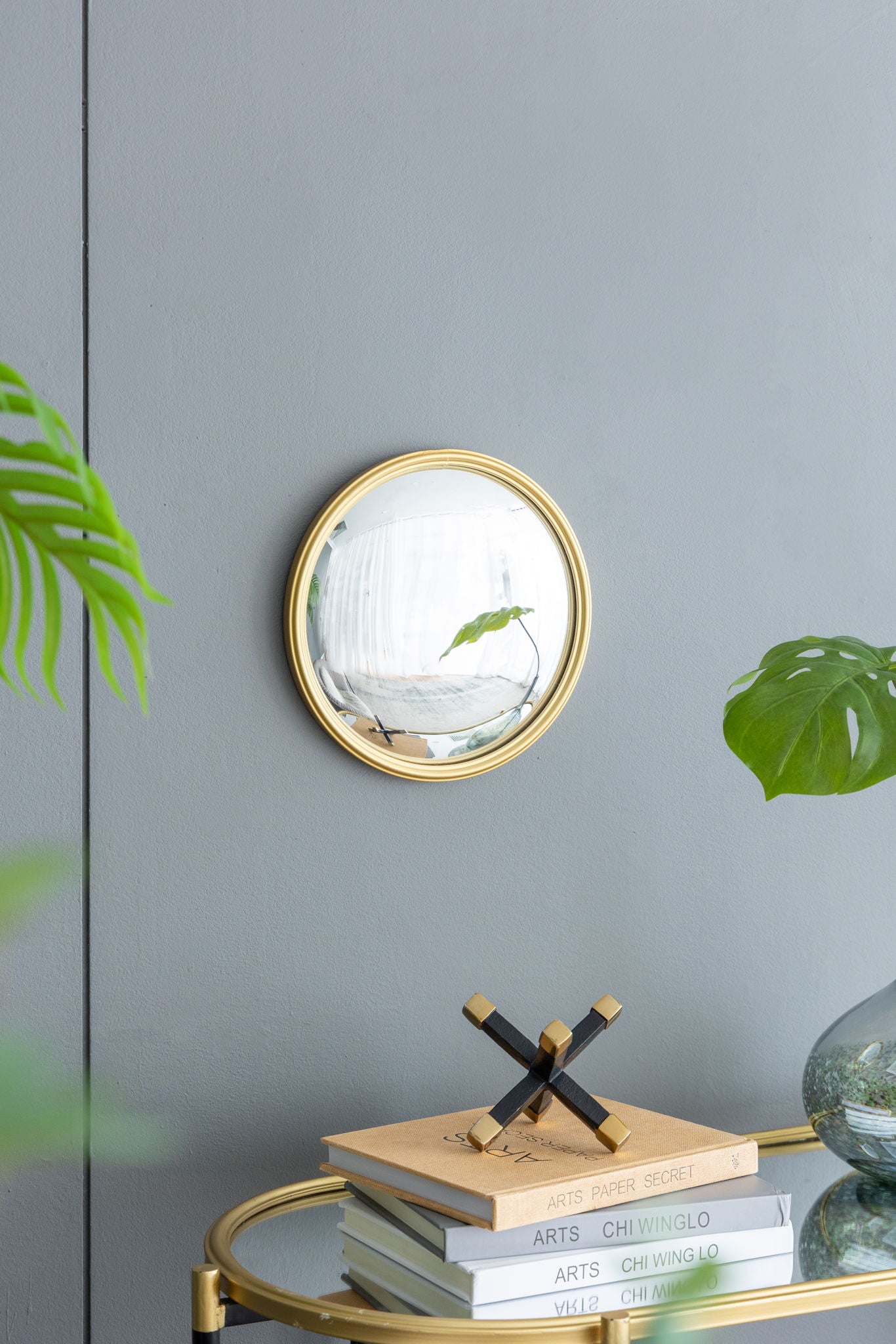 A Round Mirror, Circle Mirror With Iron Frame For Living Room Bedroom Vanity Entryway Hallway is mounted on a gray wall above a gold-trimmed table with books, a decor piece, and a vase of green leaves; a plant leaf appears at left.