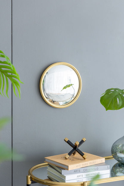 A Round Mirror, Circle Mirror With Iron Frame For Living Room Bedroom Vanity Entryway Hallway is mounted on a gray wall above a gold-trimmed table with books, a decor piece, and a vase of green leaves; a plant leaf appears at left.