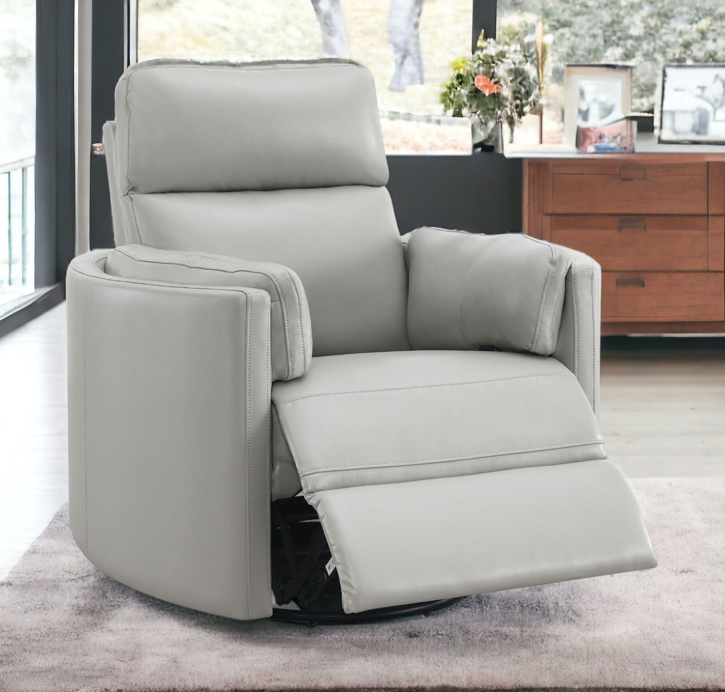 The Sagen - Chenille Glider Recliner with Swivel, in light gray, sits on a soft rug in a modern living room. Behind it are large windows and a wooden dresser adorned with framed photos and a vase of flowers, crafting a warm, cozy atmosphere.