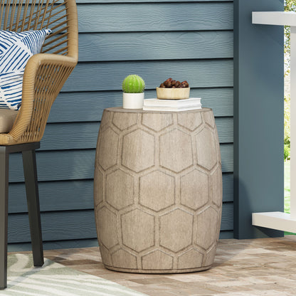 The Outdoor Lightweight Concrete Side Table - Natural, featuring a round, drum shape with a hexagonal pattern, sits on a patio beside a wicker chair. On top are a small potted cactus, a white book, and snacks. Blue siding forms the backdrop.